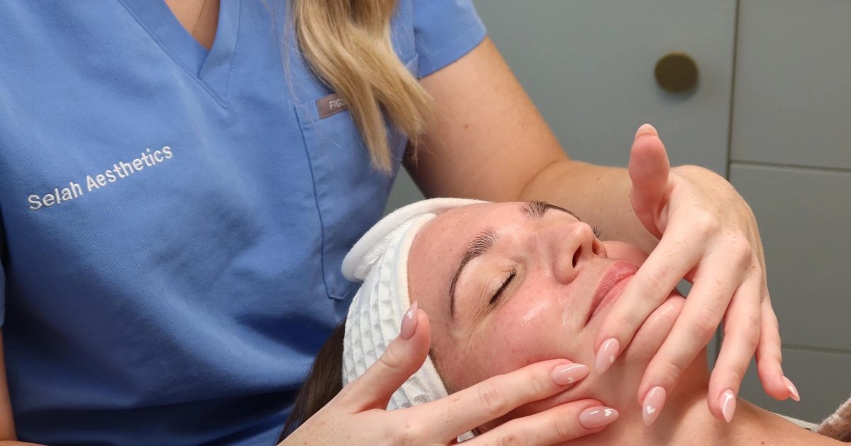 A Selah Aesthetics aesthetician in blue scrubs gives a facial massage during Skin Peel Treatments in Fairfield, AU, in a modern clinic with a marble backsplash.